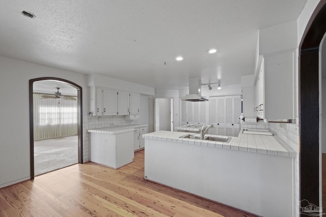 kitchen featuring kitchen peninsula, decorative backsplash, wall chimney exhaust hood, white cabinetry, and tile counters