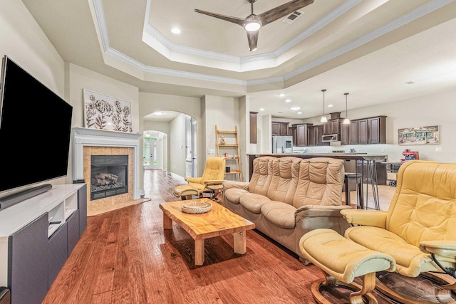 living room with ceiling fan, dark hardwood / wood-style floors, a tray ceiling, a tiled fireplace, and ornamental molding