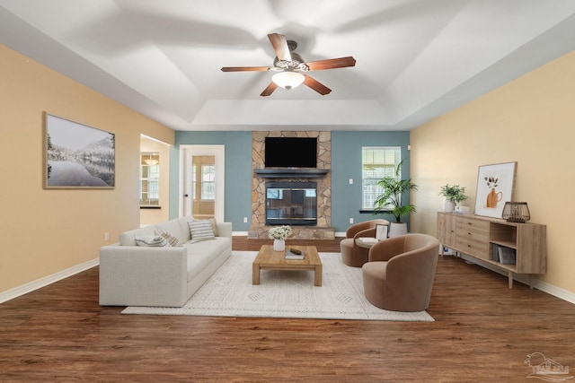 living room featuring a stone fireplace, ceiling fan, dark wood-type flooring, and a raised ceiling