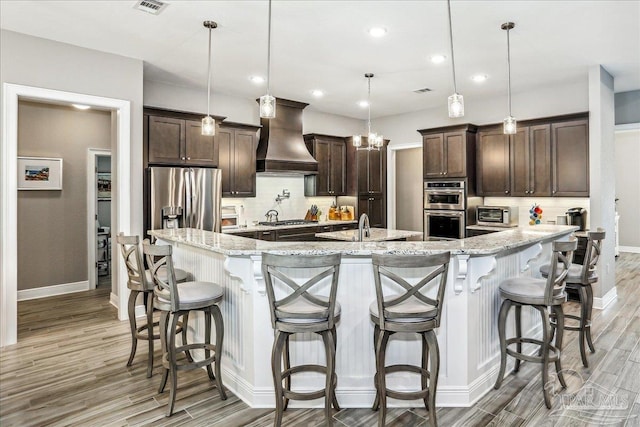 kitchen featuring custom exhaust hood, stainless steel appliances, light wood-style flooring, and dark brown cabinets