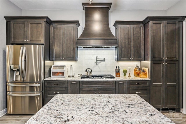 kitchen with light stone counters, dark brown cabinetry, custom range hood, wood finished floors, and appliances with stainless steel finishes