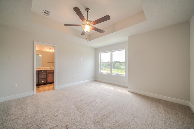 unfurnished bedroom with ceiling fan, a tray ceiling, light colored carpet, and ensuite bath