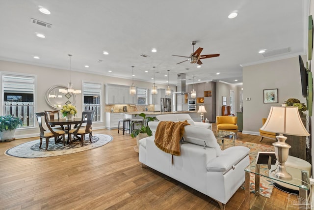 living area featuring recessed lighting, ceiling fan with notable chandelier, visible vents, light wood-style floors, and ornamental molding