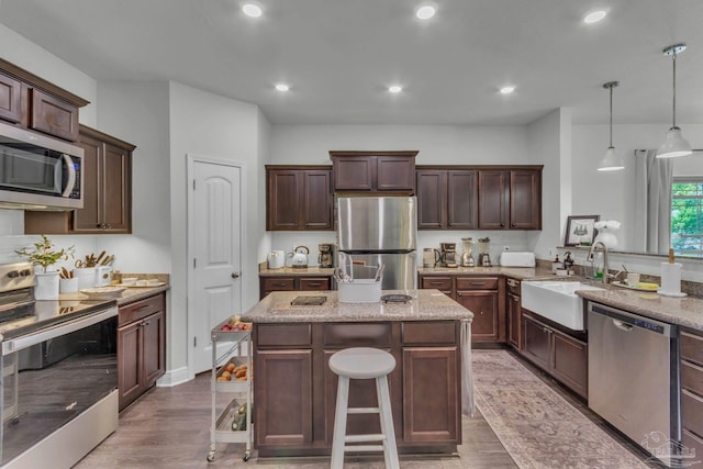 kitchen with a sink, appliances with stainless steel finishes, dark brown cabinetry, a kitchen island, and light stone counters