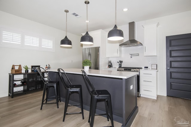 kitchen with white cabinets, wall chimney range hood, hanging light fixtures, light hardwood / wood-style flooring, and stainless steel appliances