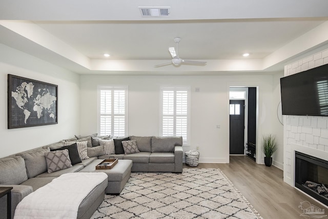 living room with ceiling fan, light wood-type flooring, a fireplace, and a tray ceiling