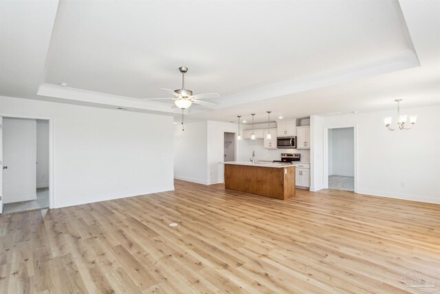 unfurnished living room with ceiling fan with notable chandelier, baseboards, a tray ceiling, and light wood-style floors