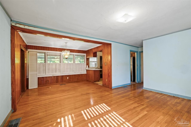 unfurnished living room with an inviting chandelier, wooden walls, light wood-type flooring, and crown molding