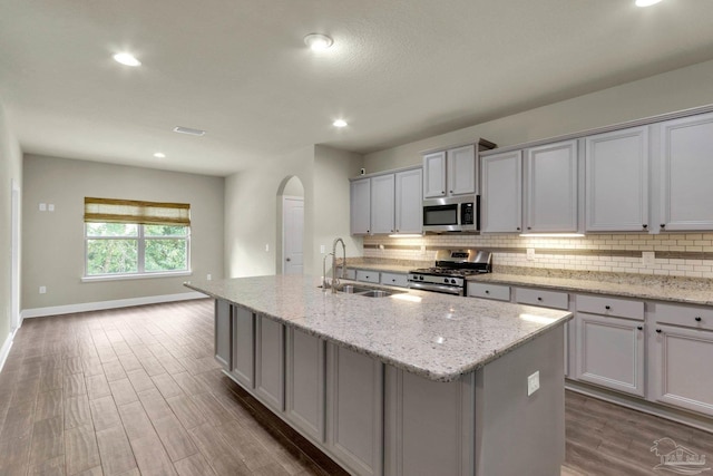 kitchen featuring light stone counters, a kitchen island with sink, appliances with stainless steel finishes, wood-type flooring, and sink