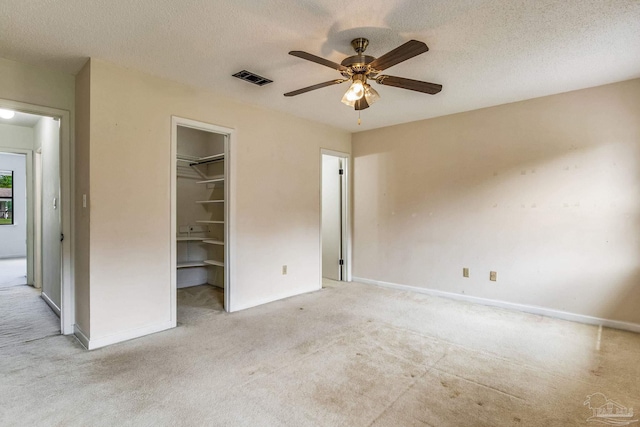 unfurnished bedroom with a walk in closet, a textured ceiling, light colored carpet, a closet, and ceiling fan