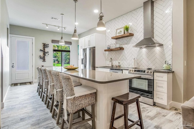 kitchen with white cabinets, light hardwood / wood-style flooring, appliances with stainless steel finishes, wall chimney range hood, and a breakfast bar area