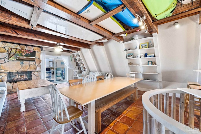 dining room featuring ceiling fan, a stone fireplace, and beamed ceiling