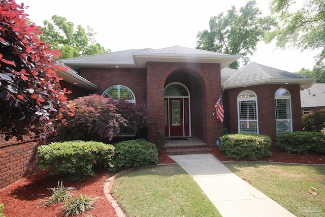 doorway to property featuring a lawn, brick siding, and roof with shingles