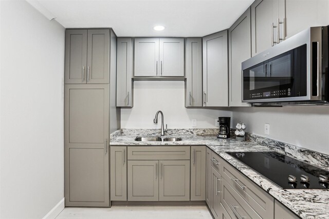 kitchen with black electric cooktop, gray cabinets, stainless steel microwave, light stone counters, and a sink