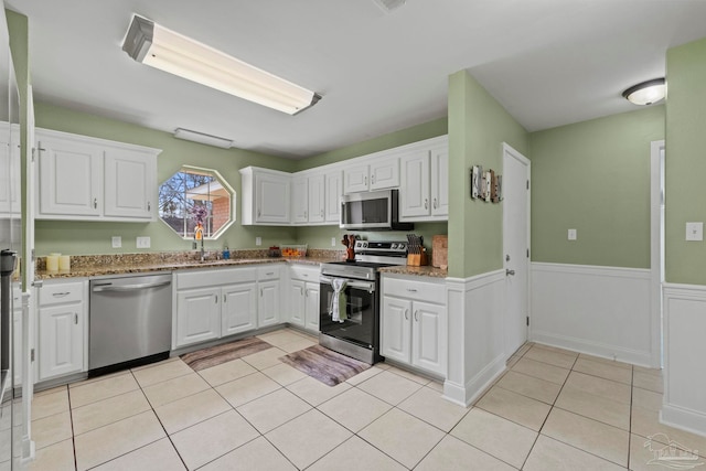 kitchen with white cabinets, wainscoting, appliances with stainless steel finishes, light stone countertops, and light tile patterned flooring