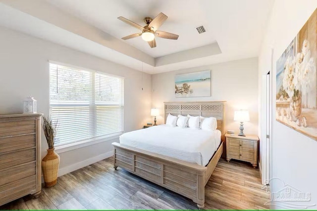 bedroom with a tray ceiling, visible vents, baseboards, wood finished floors, and ceiling fan