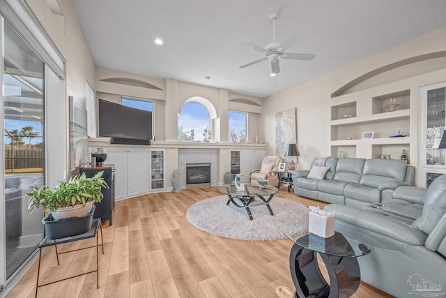 living area featuring built in shelves, a fireplace, ceiling fan, a textured ceiling, and light wood-type flooring