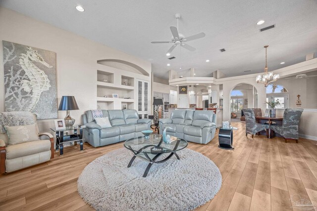 living room with arched walkways, light wood-style flooring, a textured ceiling, built in shelves, and ceiling fan with notable chandelier