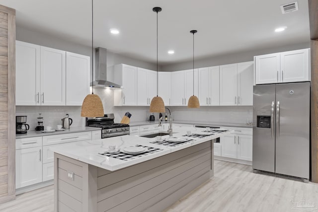 kitchen featuring white cabinetry, hanging light fixtures, wall chimney range hood, an island with sink, and appliances with stainless steel finishes