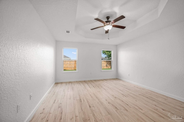 empty room with a textured wall, ceiling fan, light wood-style floors, baseboards, and a tray ceiling