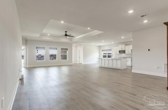 unfurnished living room with a tray ceiling, baseboards, light wood finished floors, visible vents, and a ceiling fan