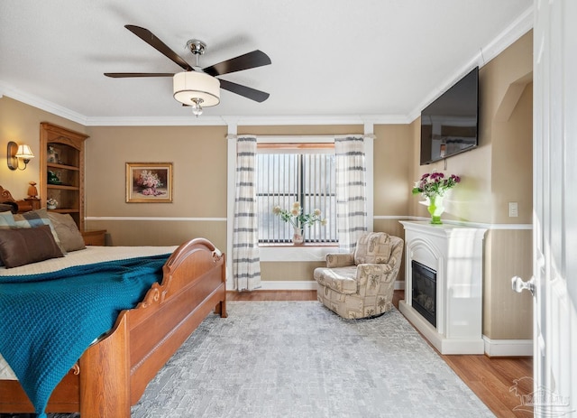 bedroom featuring crown molding, wood finished floors, a glass covered fireplace, and a ceiling fan