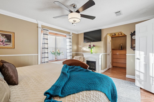 bedroom with crown molding, wood finished floors, a glass covered fireplace, a textured ceiling, and visible vents