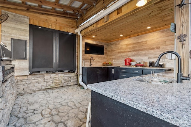 kitchen with a sink, wooden ceiling, wood walls, and light stone counters