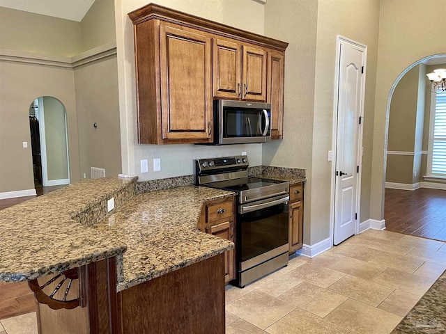 kitchen featuring arched walkways, brown cabinetry, appliances with stainless steel finishes, baseboards, and a peninsula