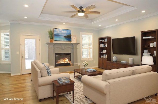 living room featuring a raised ceiling, light wood-style flooring, ornamental molding, a glass covered fireplace, and ceiling fan