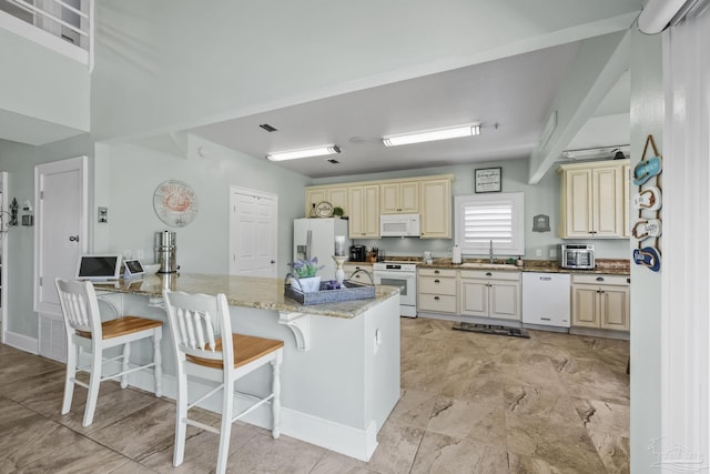 kitchen with light stone counters, white appliances, cream cabinetry, a breakfast bar area, and a sink