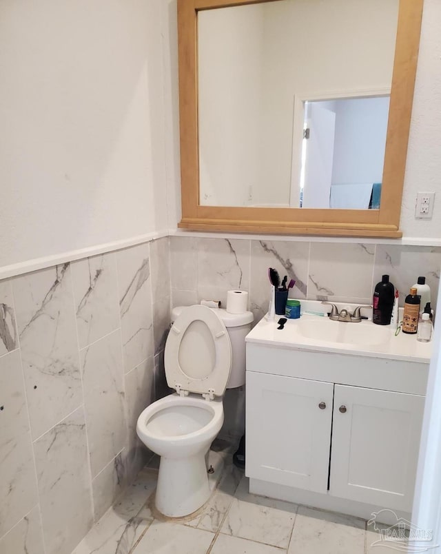 half bath featuring a wainscoted wall, toilet, vanity, marble finish floor, and tile walls