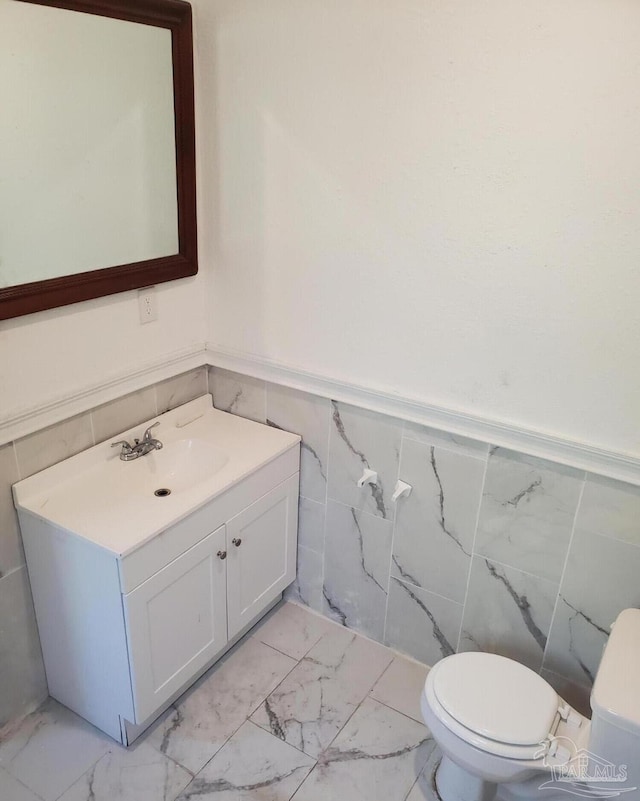 half bath with marble finish floor, toilet, vanity, and a wainscoted wall