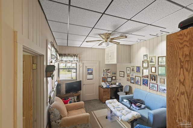 living area featuring a paneled ceiling, wooden walls, and ceiling fan