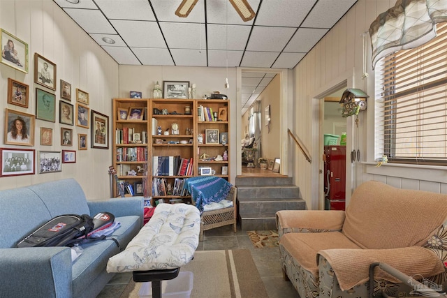 living area with stairway, a paneled ceiling, wooden walls, and ceiling fan