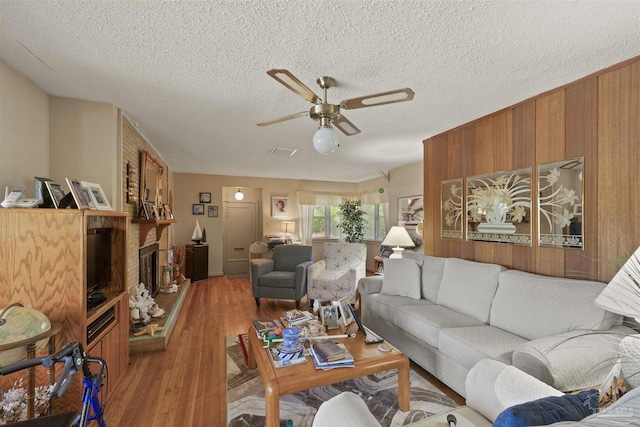 living room featuring wooden walls, light wood-style floors, a textured ceiling, a large fireplace, and ceiling fan