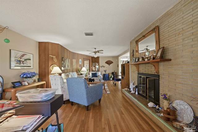 living room with a brick fireplace, wood finished floors, visible vents, a textured ceiling, and ceiling fan
