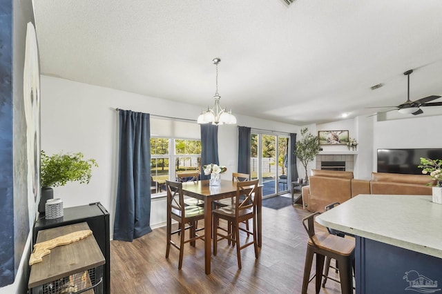 dining space with a tile fireplace, a textured ceiling, ceiling fan with notable chandelier, and dark wood-type flooring