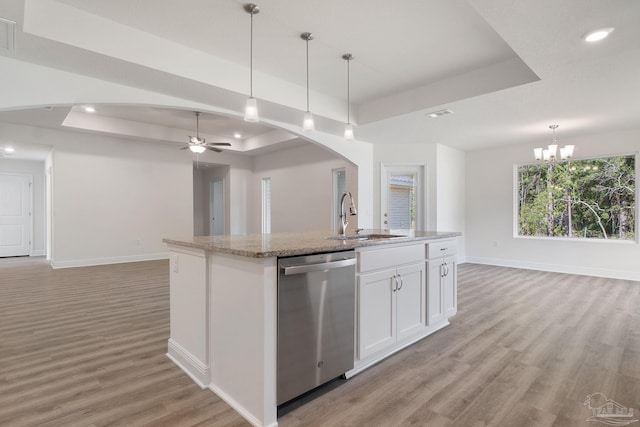 kitchen with a raised ceiling, ceiling fan with notable chandelier, an island with sink, stainless steel dishwasher, and white cabinetry