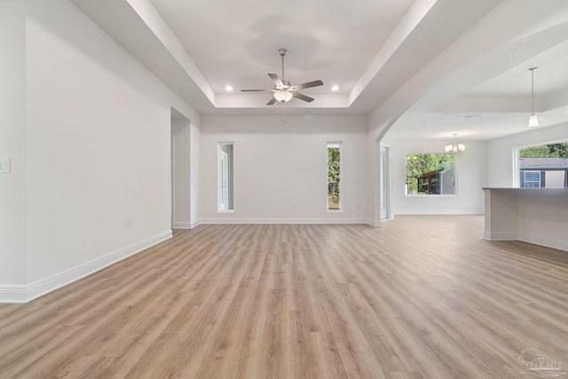 unfurnished living room with plenty of natural light, ceiling fan with notable chandelier, light wood-type flooring, and a tray ceiling