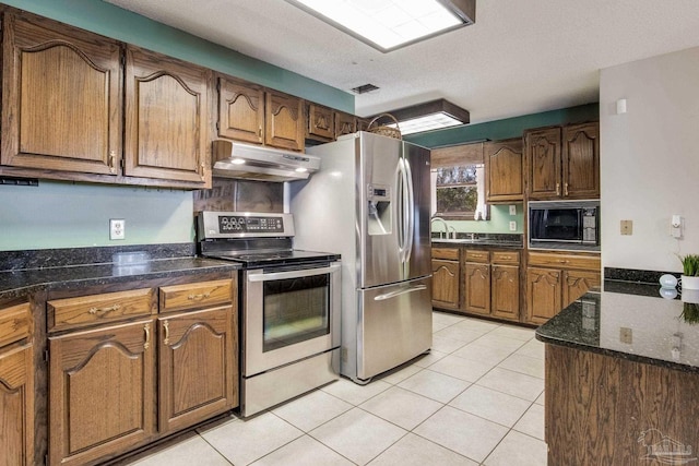 kitchen with under cabinet range hood, light tile patterned floors, brown cabinets, appliances with stainless steel finishes, and a sink