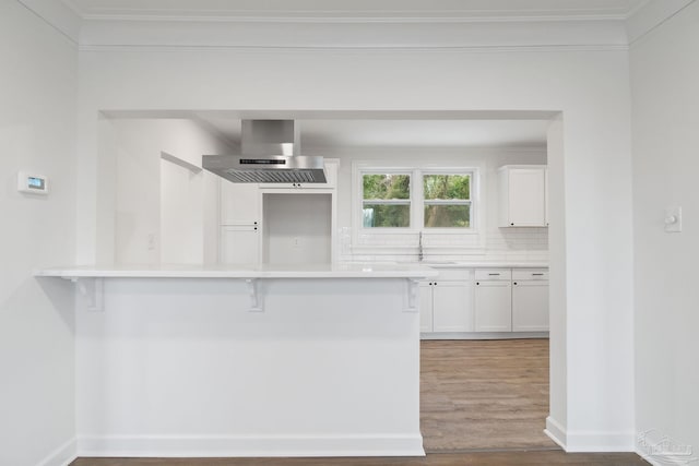 kitchen featuring wood finished floors, white cabinetry, a kitchen breakfast bar, wall chimney exhaust hood, and backsplash