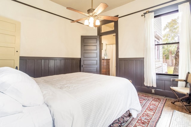 bedroom featuring multiple windows, wainscoting, wood-type flooring, and a ceiling fan