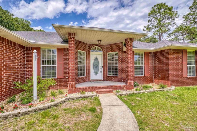 doorway to property featuring brick siding, a yard, and roof with shingles