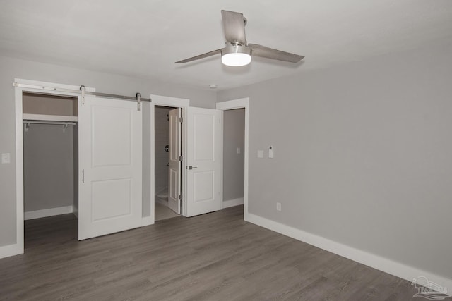 unfurnished bedroom featuring ceiling fan, a spacious closet, a barn door, dark hardwood / wood-style floors, and a closet