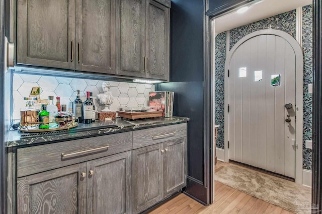 bar with dark brown cabinets, dark stone countertops, light wood-type flooring, and backsplash