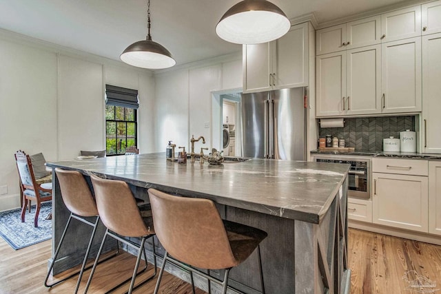 kitchen with backsplash, light wood-type flooring, an island with sink, decorative light fixtures, and stainless steel appliances