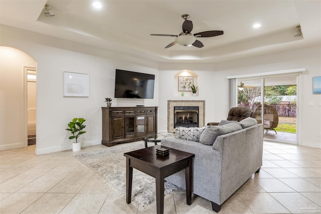 living room with ceiling fan, a raised ceiling, a tile fireplace, and light tile patterned floors