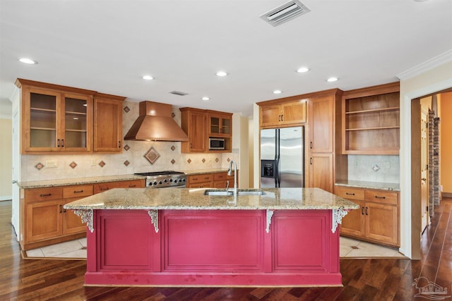 kitchen with wall chimney range hood, sink, stainless steel appliances, a kitchen breakfast bar, and a center island with sink