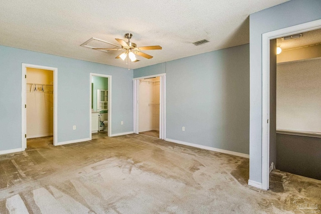 unfurnished bedroom with a spacious closet, a textured ceiling, ensuite bath, light colored carpet, and ceiling fan